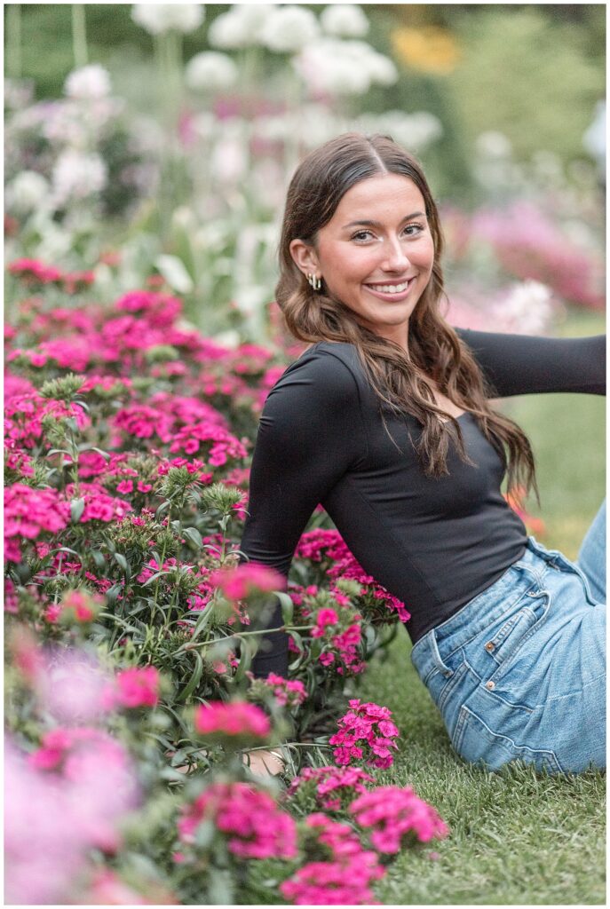 senior girl in black top and blue jeans sitting in grass by pink bush at longwood gardens in pennsylvania