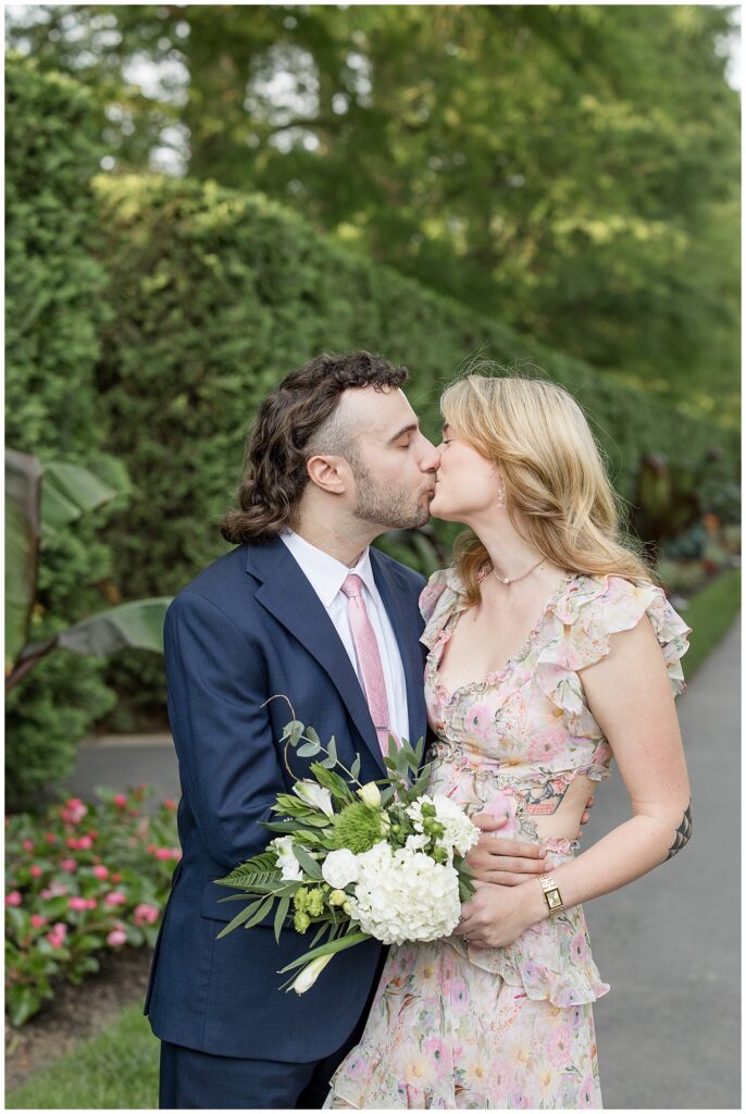 engaged couple kissing on path at longwood gardens as they hug and she holds bouquet of flowers in kennett square