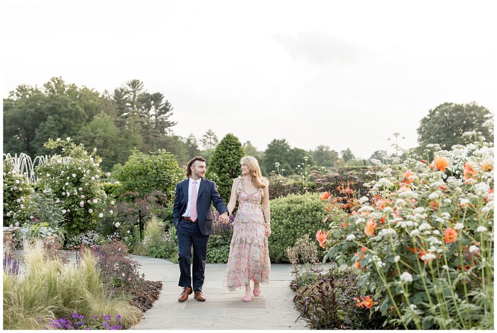 engaged couple holding hands and walking on landscaped path towards camera at longwood gardens