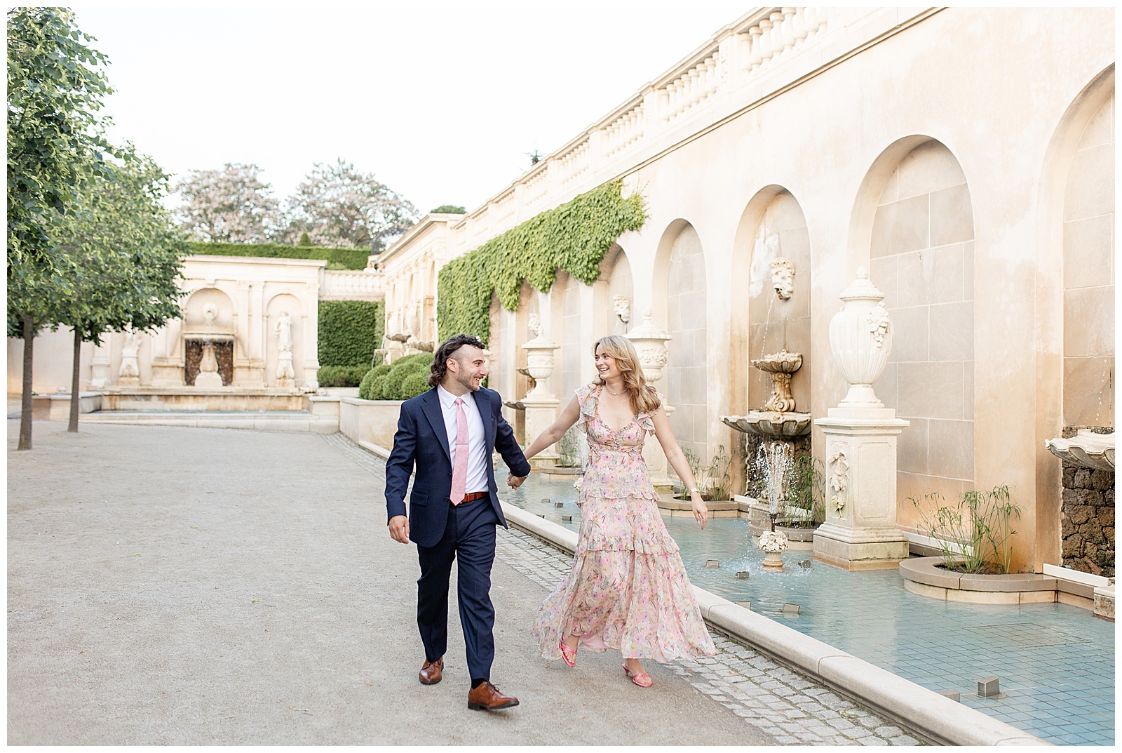 guy in navy suit with pink tie and girl in light pink flowy dress holding hands by fountains at longwood gardens