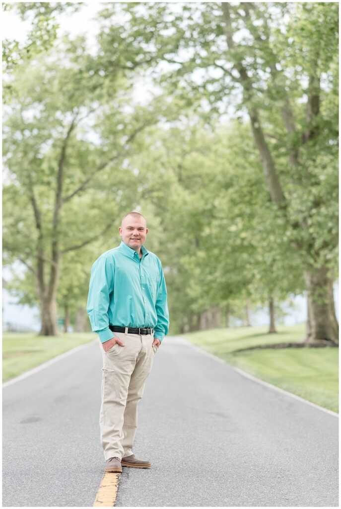 senior guy in teal dress shirt and khaki pants standing in middle of road with hands in his pockets at masonic village