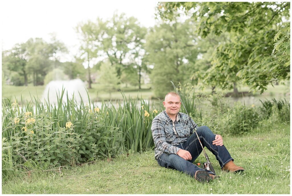 senior guy in flannel shirt and jeans sitting on grass by pond with fishing rod at masonic village