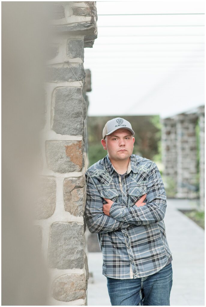 close up photo of senior guy leaning against stone column at masonic village wearing flannel shirt and baseball cap