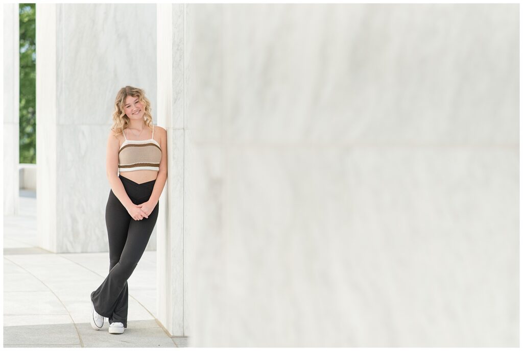 senior girl in tube top and black flare pants leaning against white concrete wall at founders hall in hershey pennsylvania