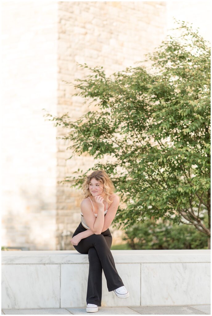 senior girl sitting on concrete ledge resting her chin on her right elbow on her knee at founders hall at milton hershey school