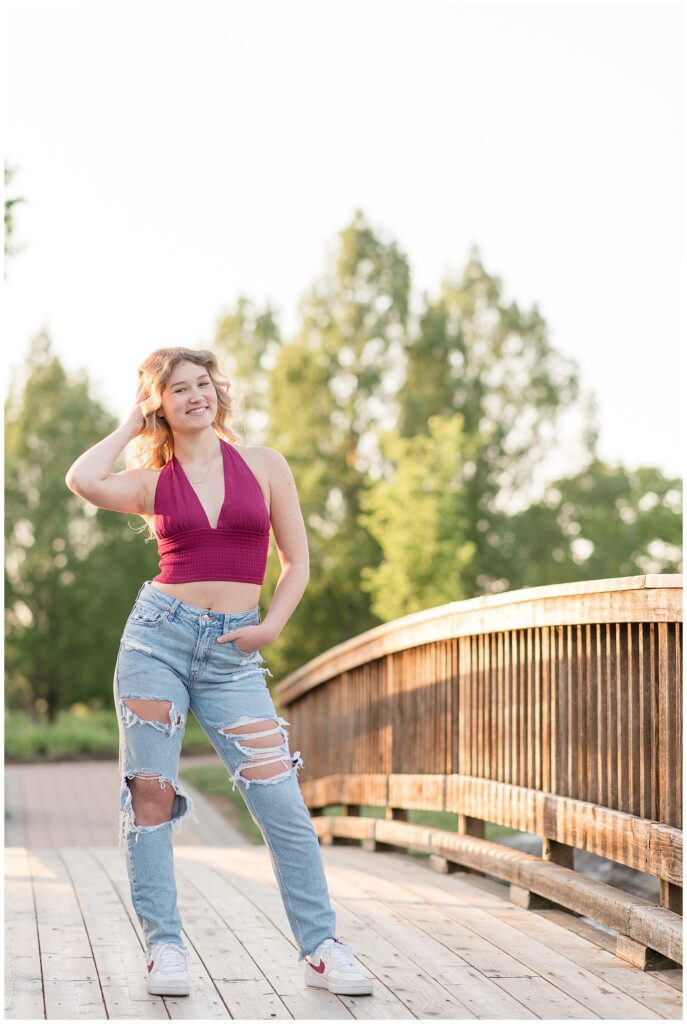 senior girl wearing maroon v-neck tank top and ripped blue jeans with her left hand on her hip on wooden bridge at sunset in hershey pa