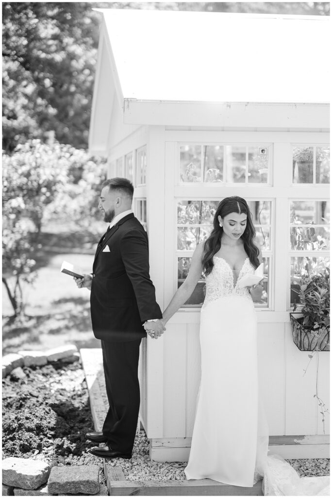 black and white photo of bride and groom holding hands around corner of white building and reading notes to each other at domain pterion