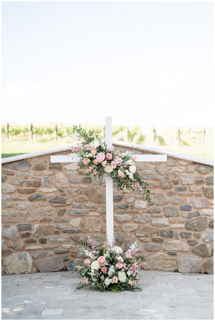 photo of large white cross with flowers on it by stone wall at domain pterion