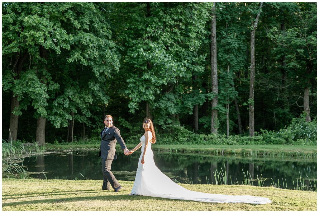 couple holding hands and walking along pond with bride's long dress train trailing behind her