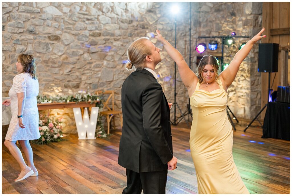 wedding guests dancing with woman raising her arms above her head inside barn at domain pterion