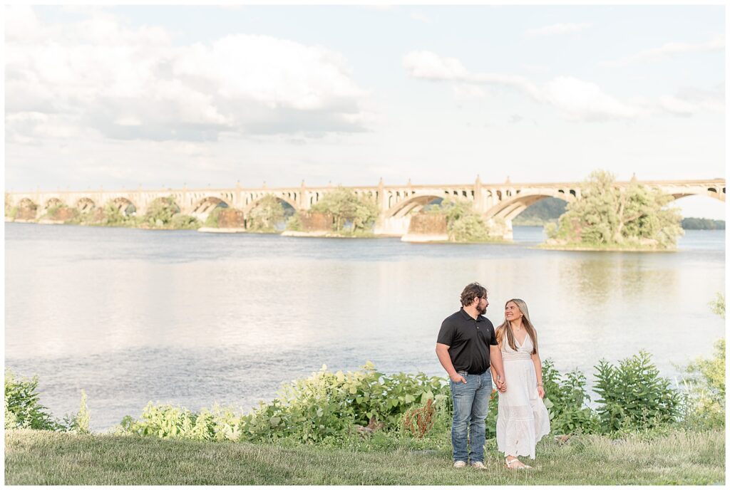 couple holding hands and looking at each other by susquehanna river with old bridge behind them in york county pennsylvania