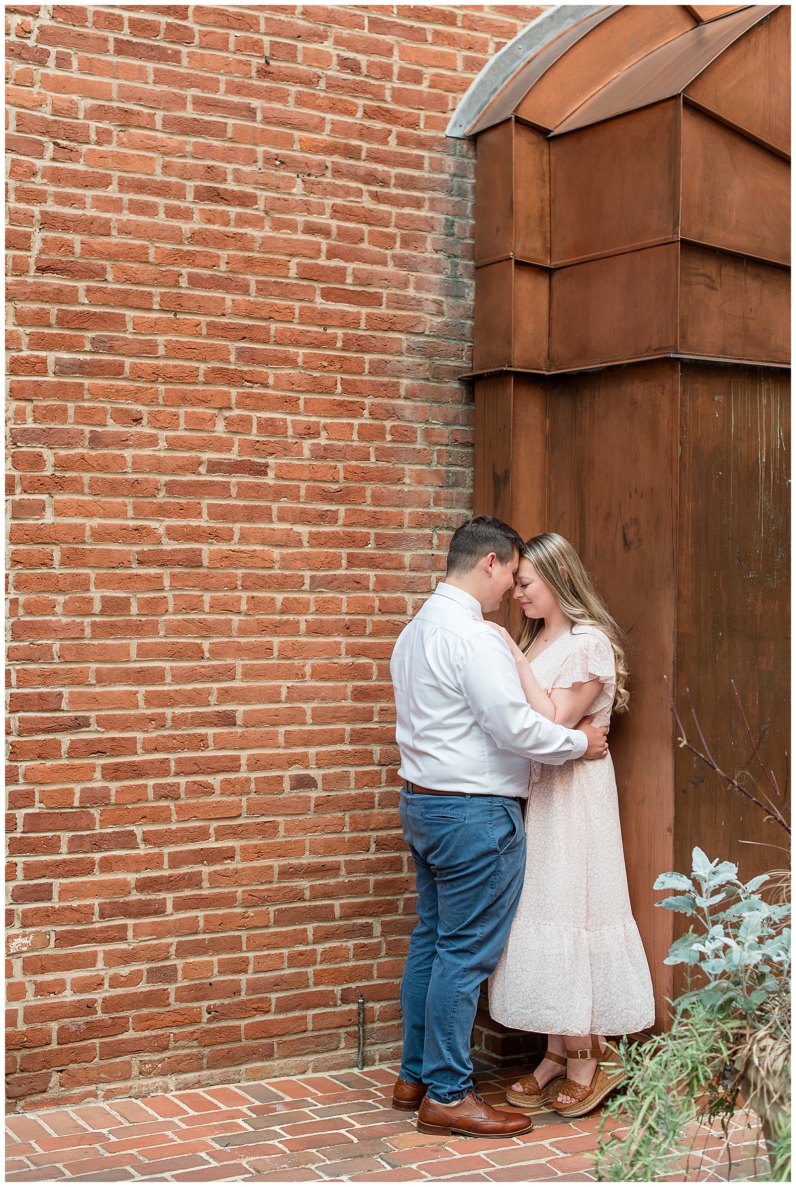 man in dress shirt and navy pants and woman in light flowy dress hugging by brick wall in lancaster city