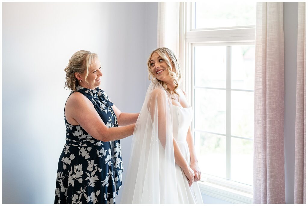 bride looking back over her right shoulder and smiling at her mom who is buttoning her gown at ironspire complex