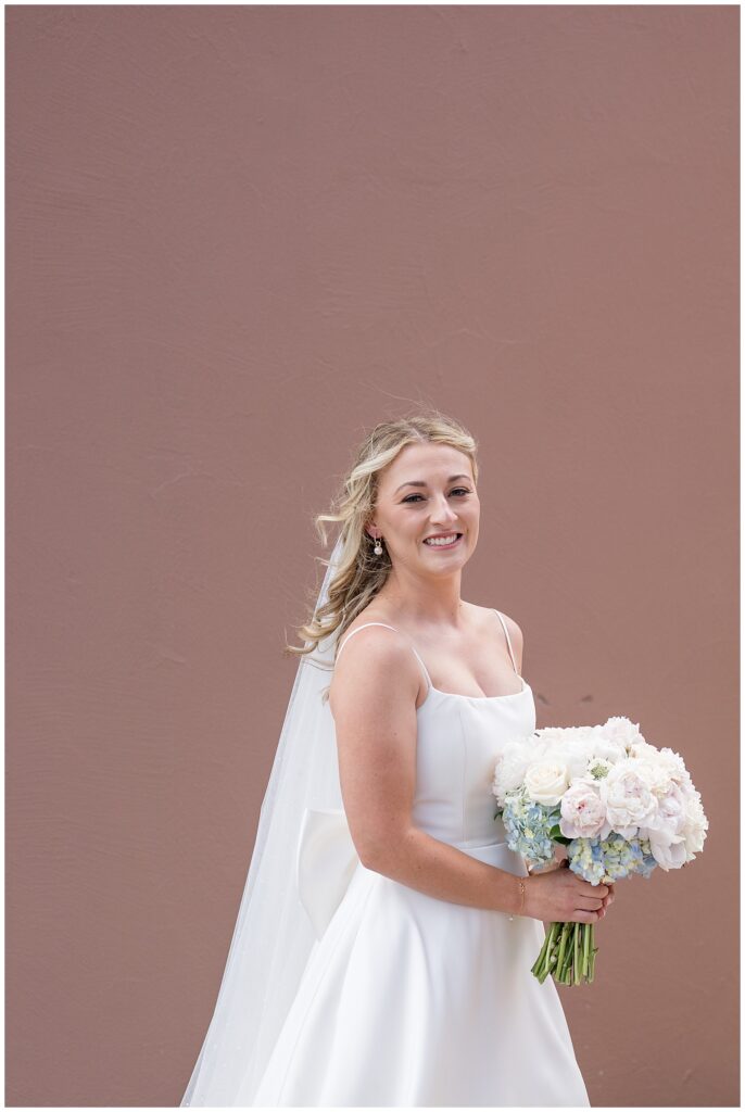 bride wearing spaghetti strap white gown with long veil and holding bouquet of white flowers by mauve wall at ironspire complex