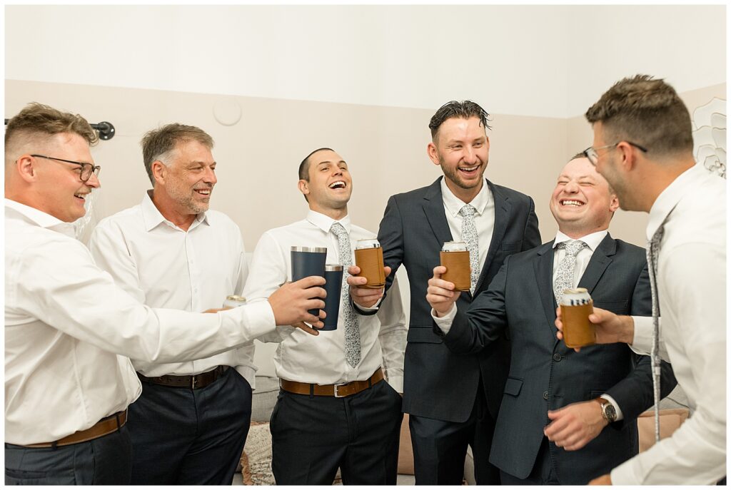 groom surrounded by his bridal party and laughing as they all hold glasses of beer at ironspire complex in lancaster county