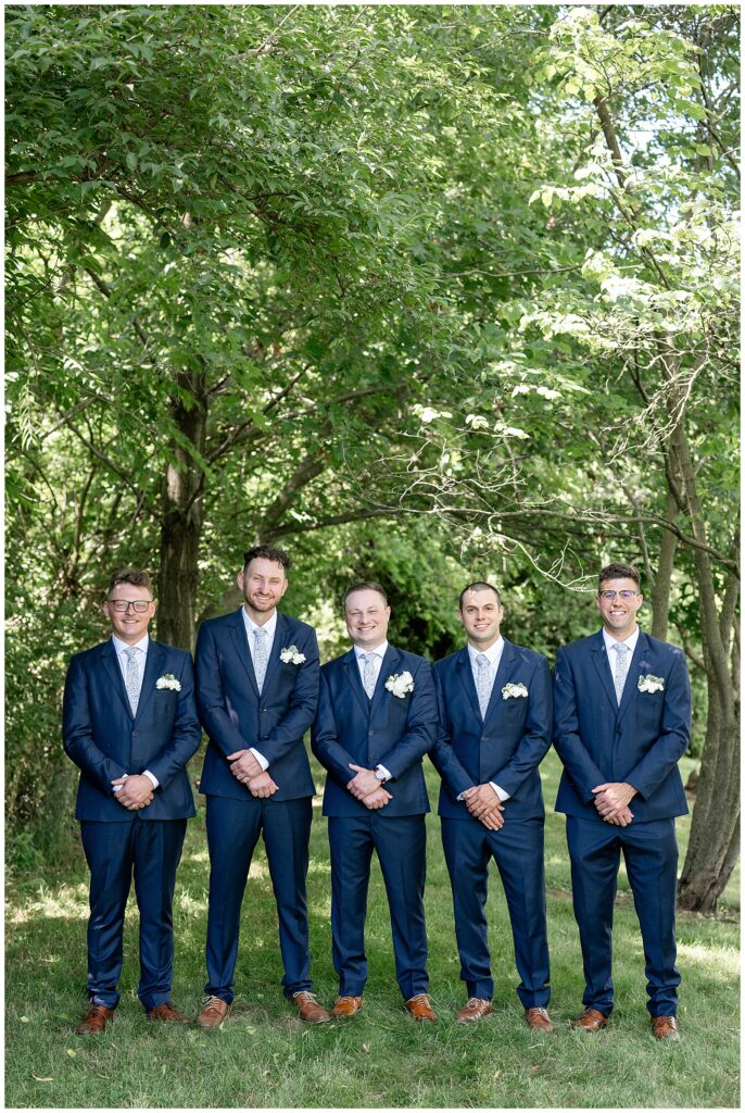 groom with his four groomsmen all wearing navy suits in wooded area at ironspire complex