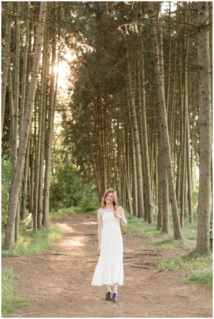 senior girl in long white dress with spaghetti straps standing in between rows of evergreen trees at overlook park