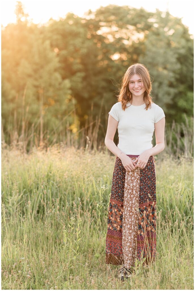 senior girl in white t-shirt and long floral skirt standing by tall wild grasses at sunset at park in lancaster county