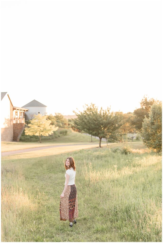 senior girl walking away from camera holding edge of long skirt at sunset with building in background at overlook park