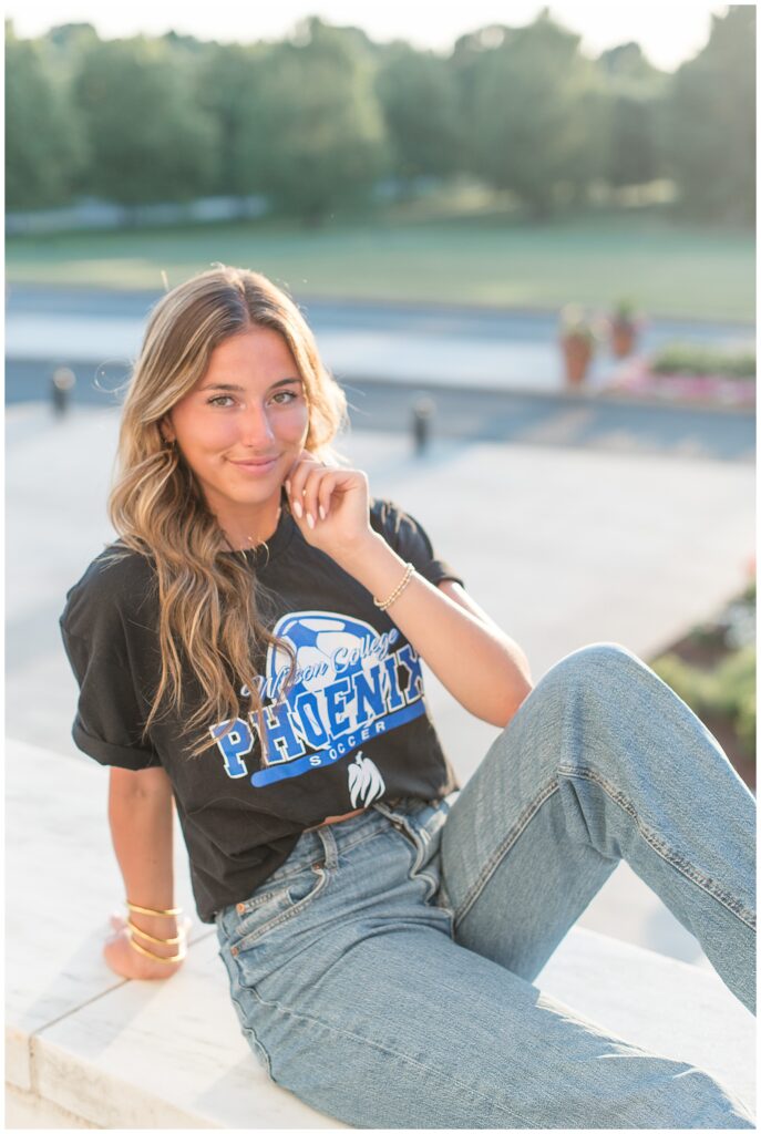 senior girl sitting on ground with left knee bent wearing college t-shirt and jeans at founders hall at milton hershey school