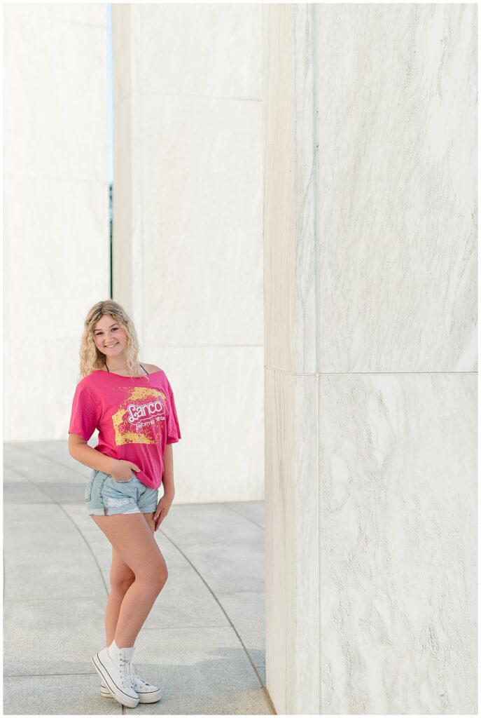 senior girl with right hand on hip and right knee popped wearing college t-shirt and shorts by concrete wall at founders hall