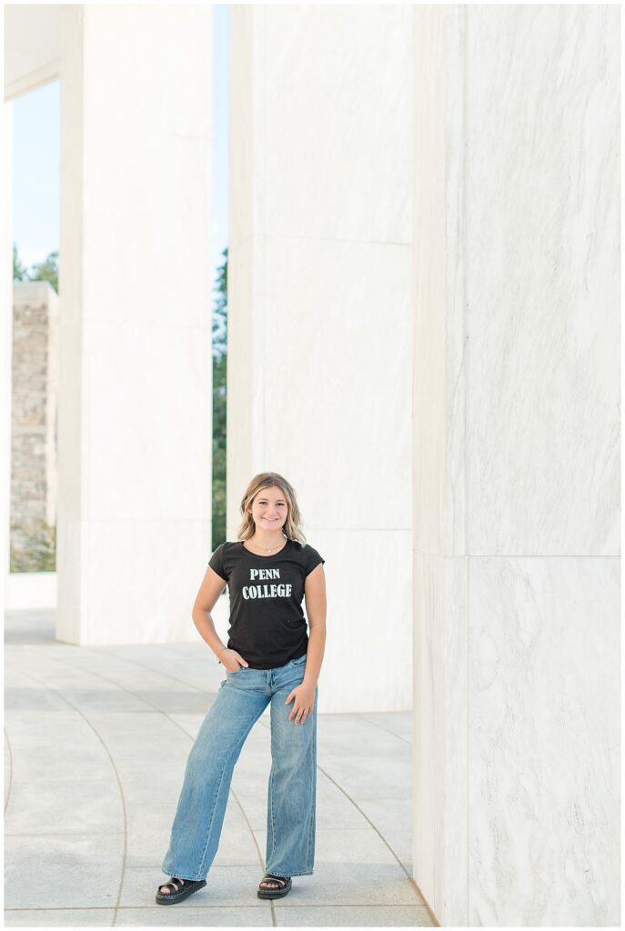 senior girl with right hand on hip by tall concrete wall wearing college t-shirt and jeans at founder's hall