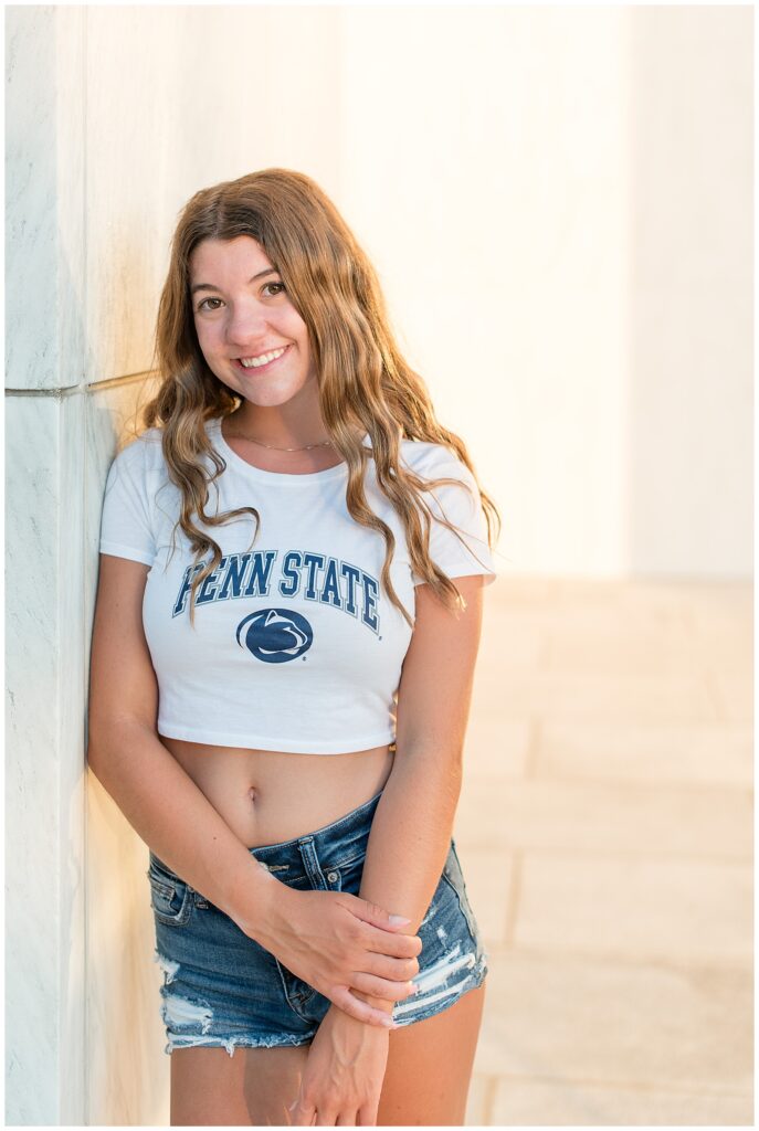 senior girl leaning right shoulder against concrete wall wearing penn state crop top and jean shorts at milton hershey school