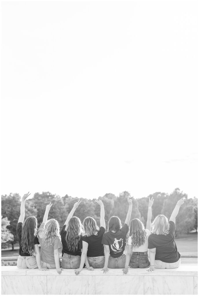 black and white photo of senior girls sitting on wall with backs to camera and all waving hand in the air at founders hall