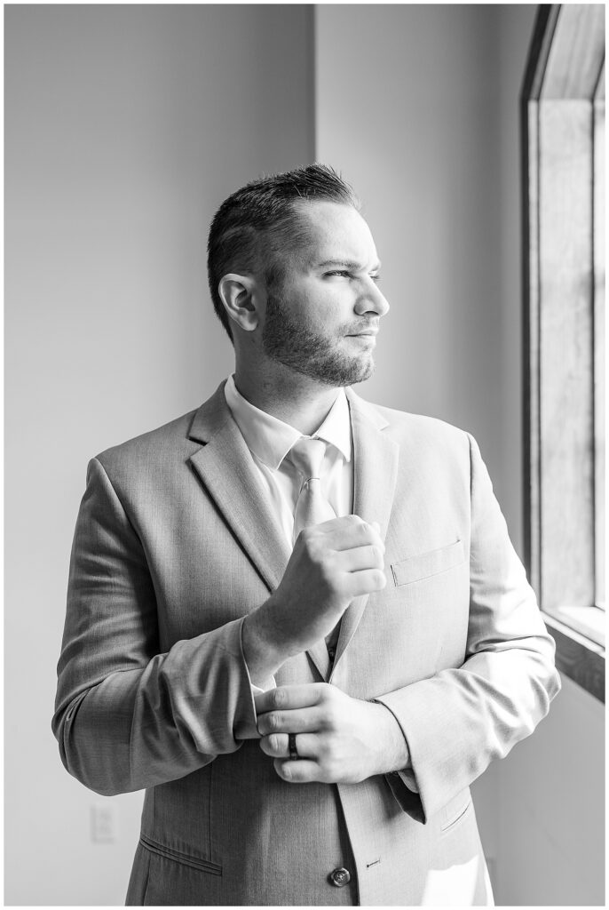 black and white photo of groom looking left out window as he adjusts his suit sleeve at folino estate winery