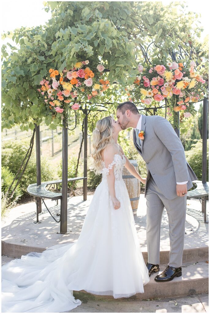 bride and groom sharing their first kiss by archway covered in colorful flowers at folino estate