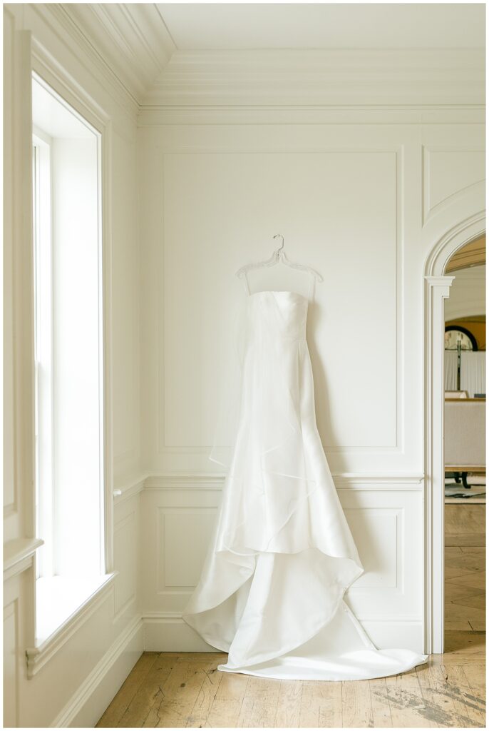 elegant and simple wedding dress hanging up against a white wall with beautiful trim