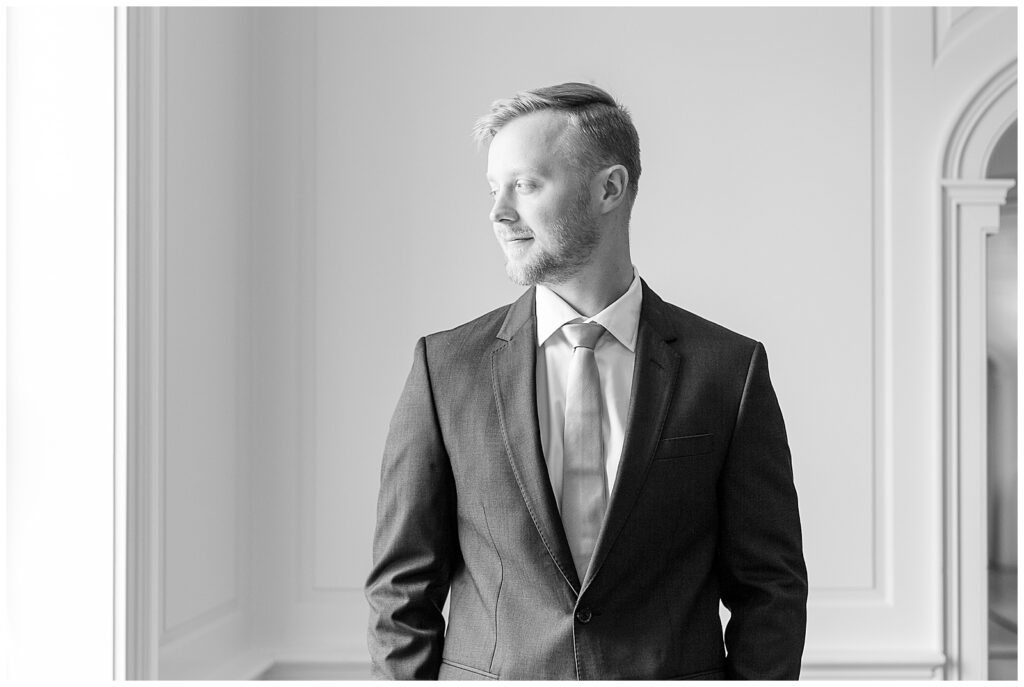 black and white groom portrait of him looking to side out window with hands in his pockets