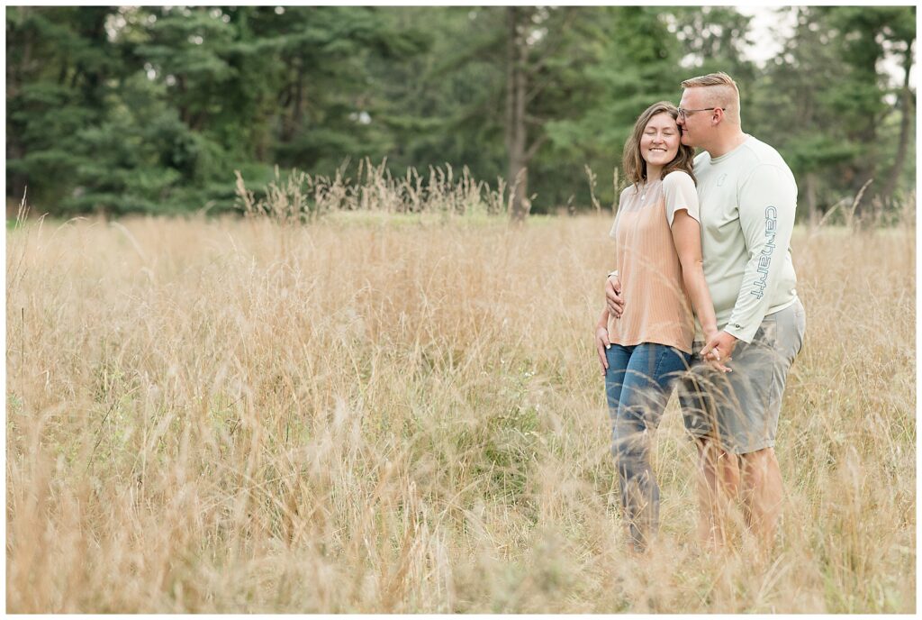 guy standing behind girl as they hold hands and he kisses her in field of dried wild grasses at lancaster central park