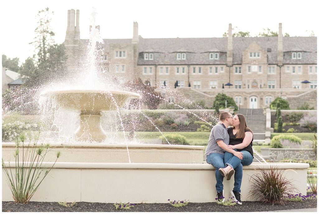 engaged couple sitting on fountain ledge and kissing with large building behind them in elizabethtown pa