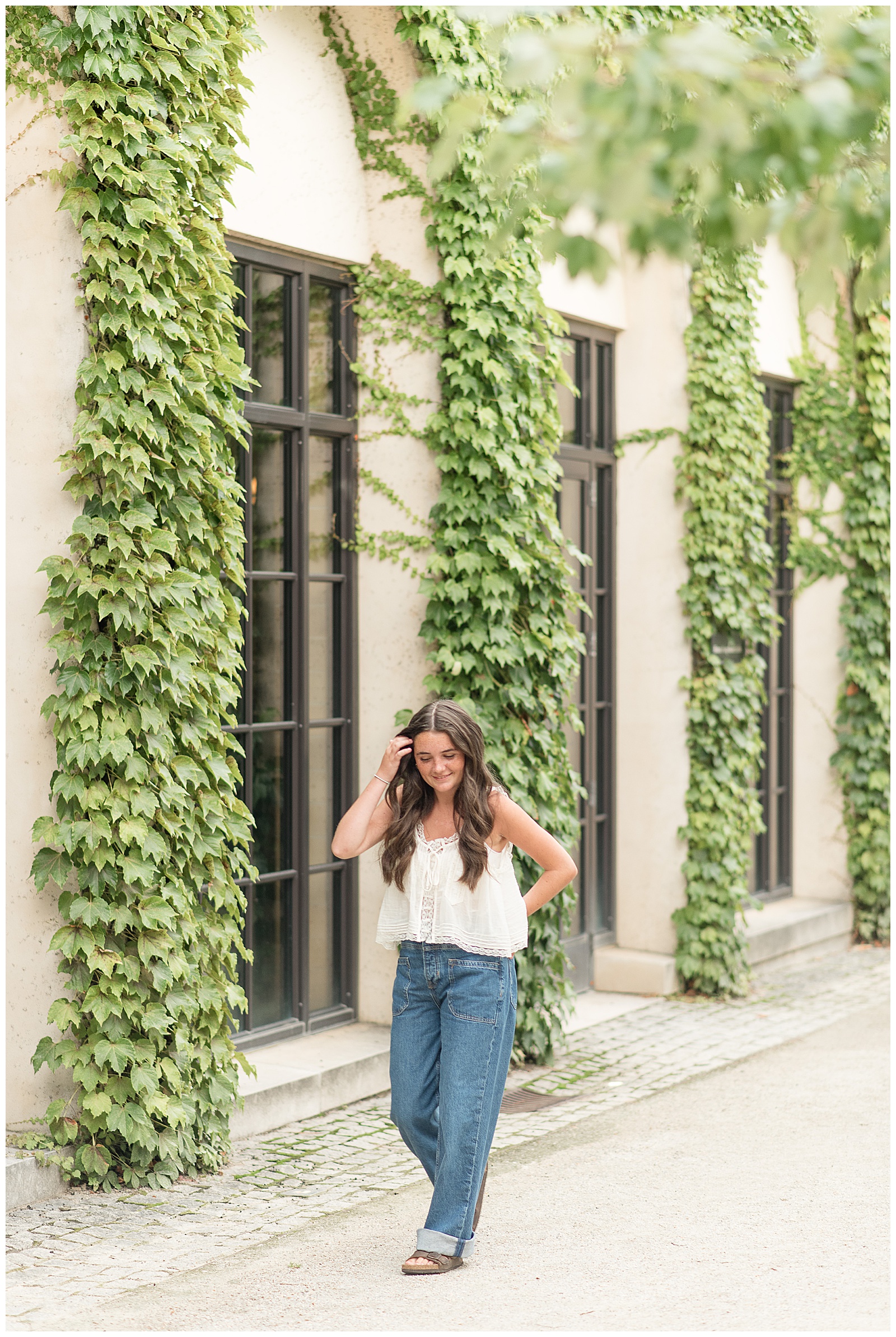 senior girl in white tank top and jeans tucking long hair behind her right ear by ivy wall at longwood gardens