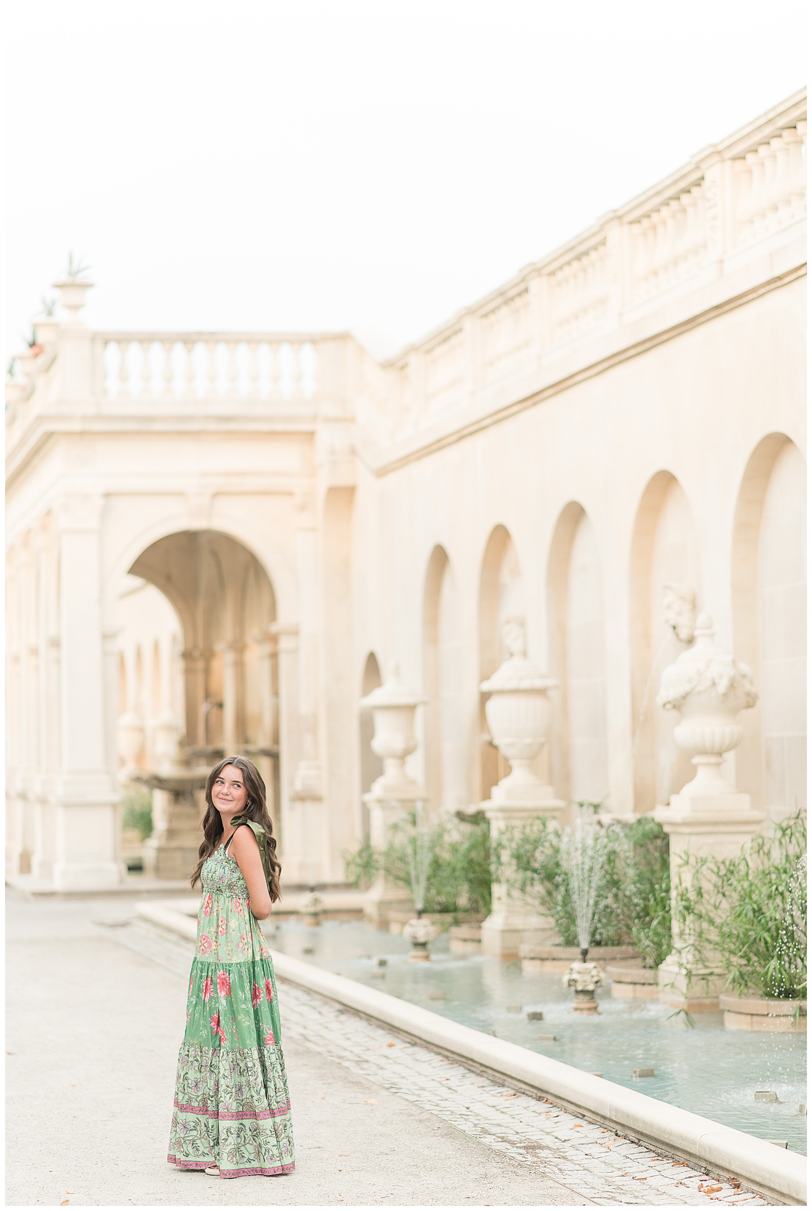 senior girl in sage green floral dress walking away from camera looking back over left shoulder by fountains at longwood gardens