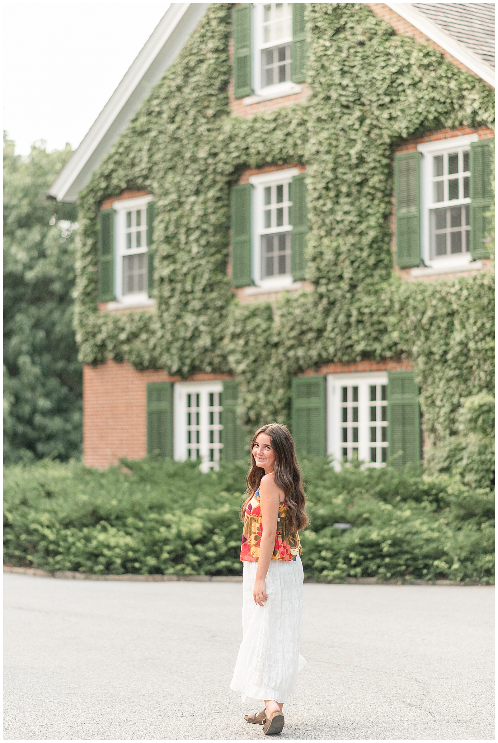 senior girl walking away from camera looking back over left shoulder by ivy covered brick house at longwood gardens