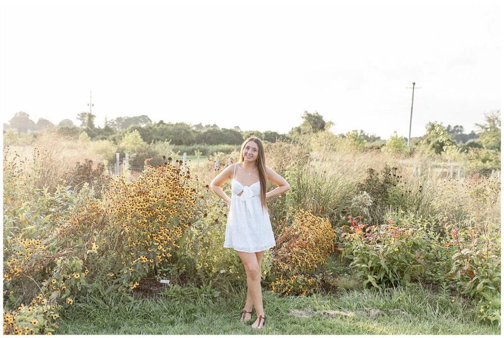 senior girl with hands on hips wearing cute white dress in field of wildflowers at overlook park