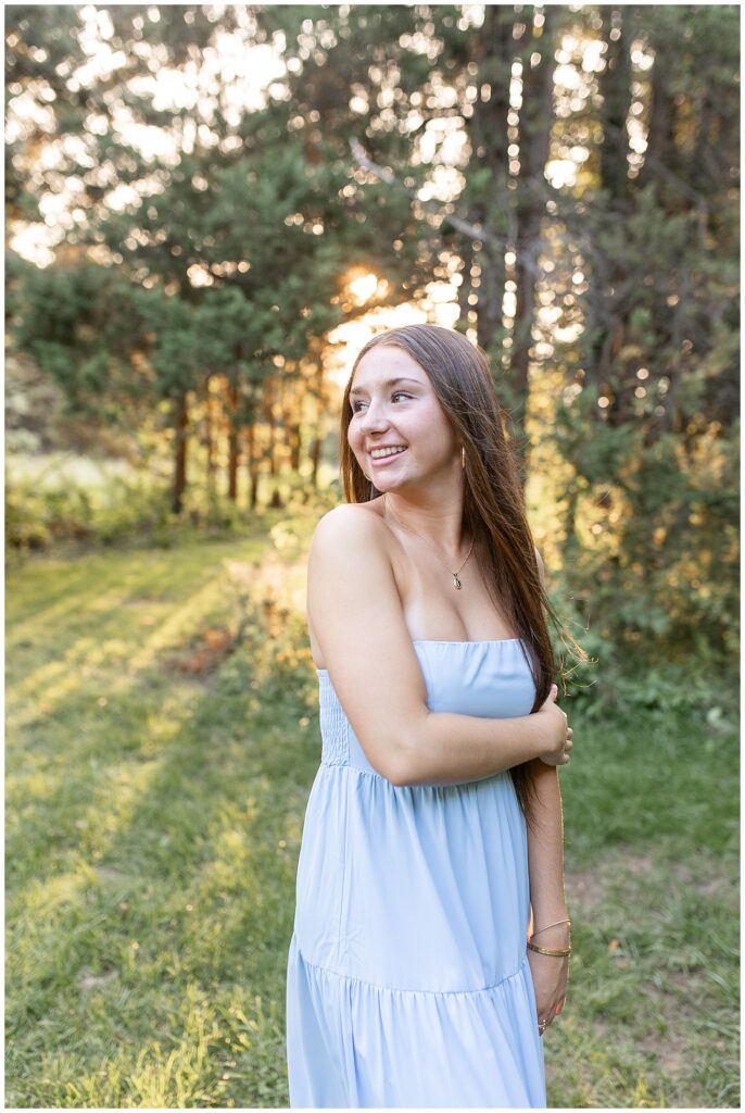 senior girl with right shoulder toward camera by rows of evergreen trees with sunset peeking through behind her in lancaster pennsylvania