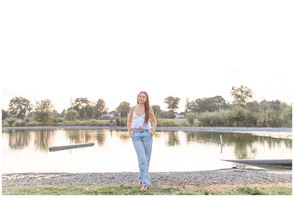 senior girl in white tank top and blue jeans with hands in pockets by pond at overlook park