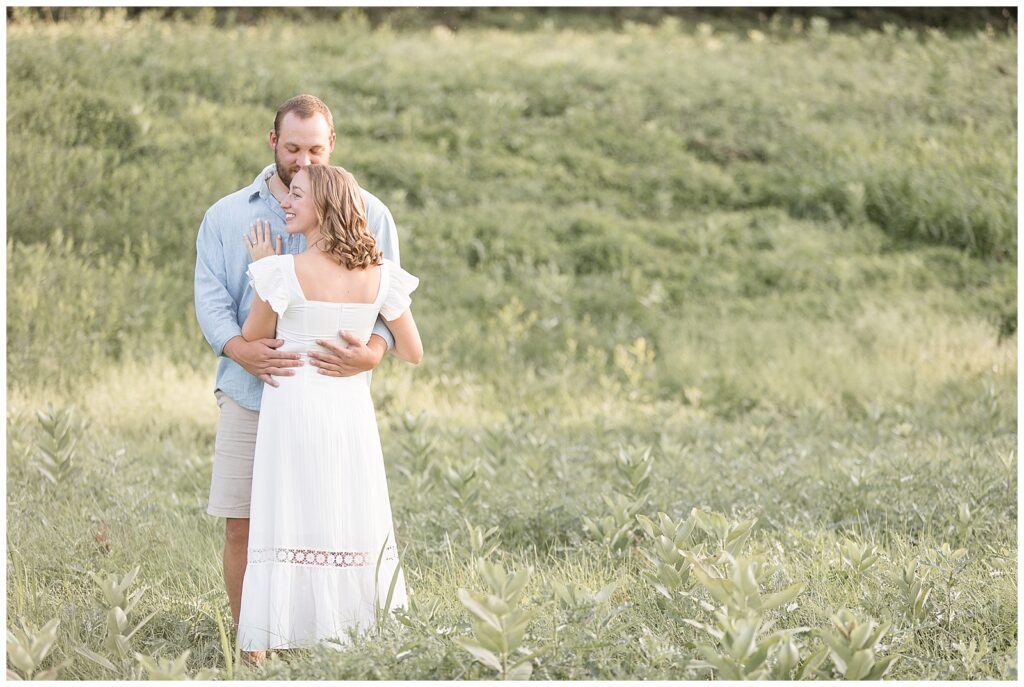 engaged couple hugging with woman's back to camera as she looks to her left and man looks smiling at her in field at hibernia park