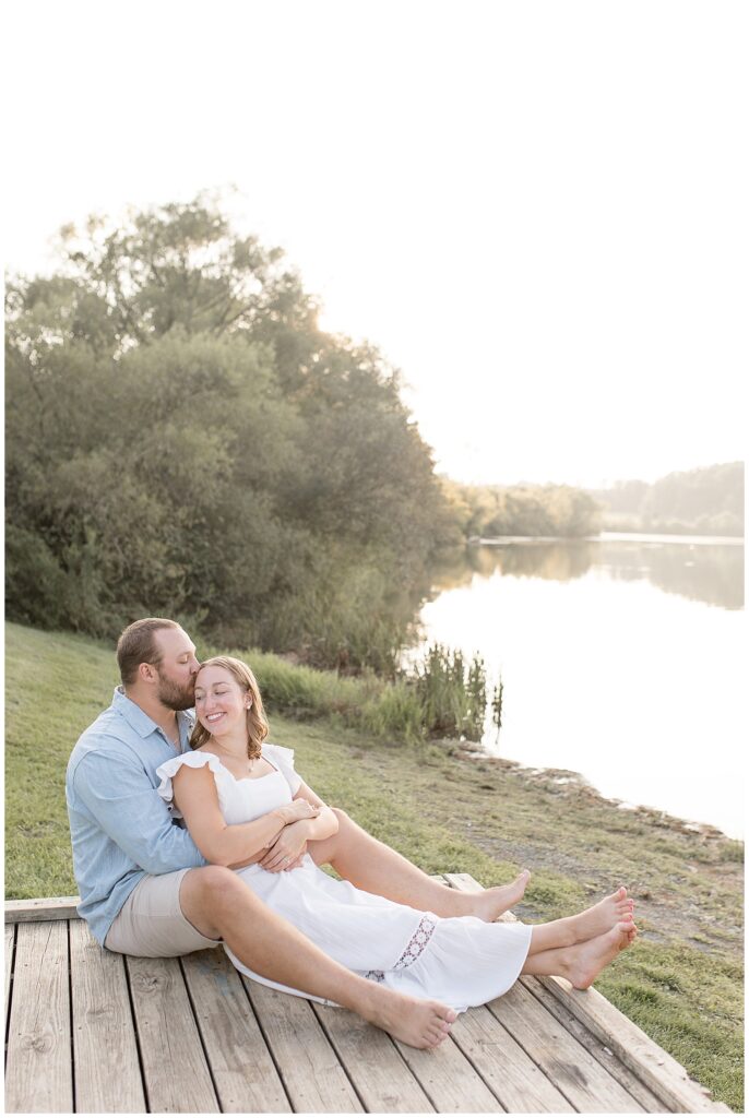 man sitting behind woman as they both sit on wooden dock and he kisses her forehead by lake at hibernia park