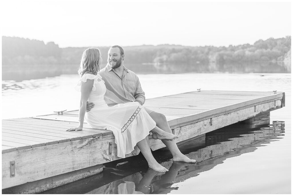 black and white photo of couple sitting on dock and smiling at each other at hibernia park