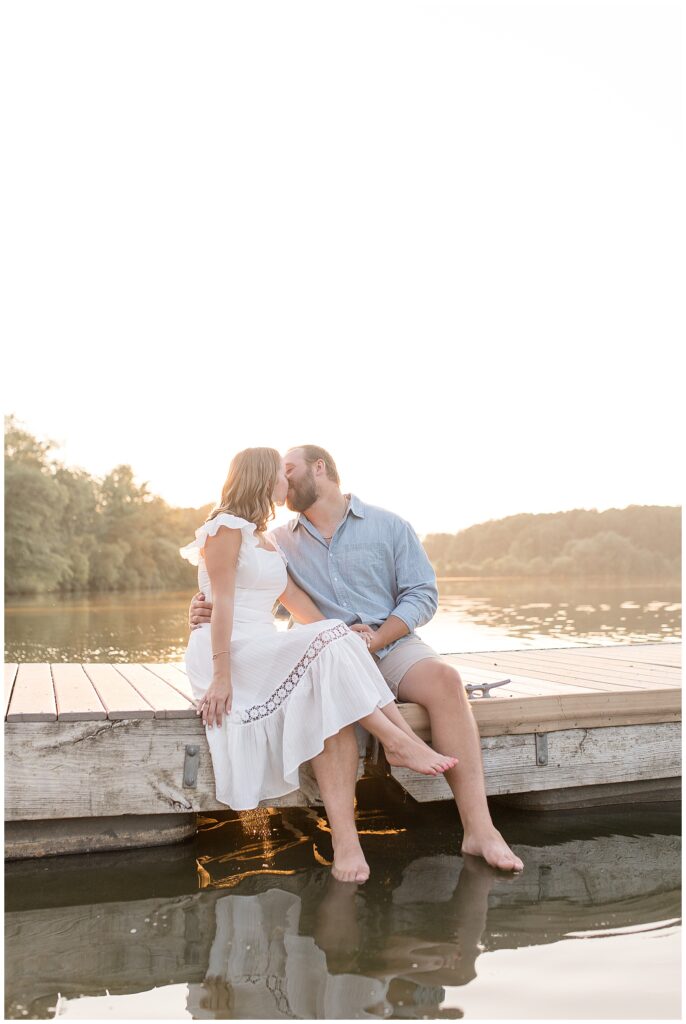 engaged couple sitting on dock kissing by lake at sunset at hibernia park