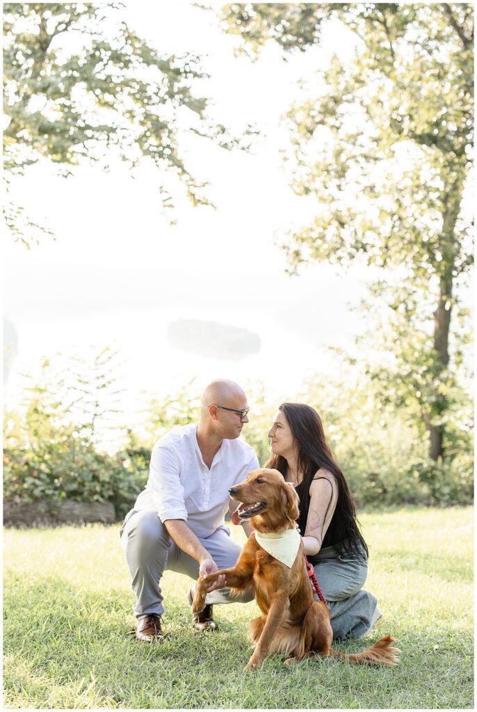 engagement couple crouching down with their pup and looking at each other on sunny night at pinnacle point