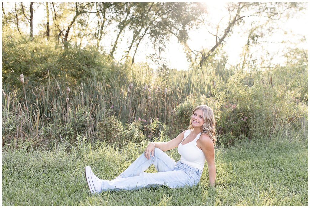 senior girl in white tank top and blue jeans sitting in the grass with sun shining through the trees at overlook park