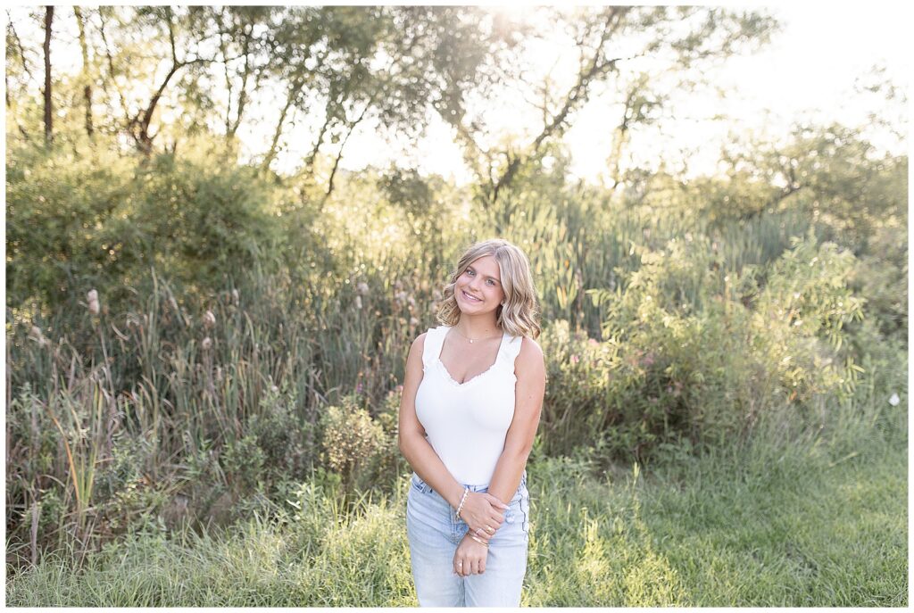 senior girl smiling at camera with right hand holding left wrist at sunset at park in lancaster county