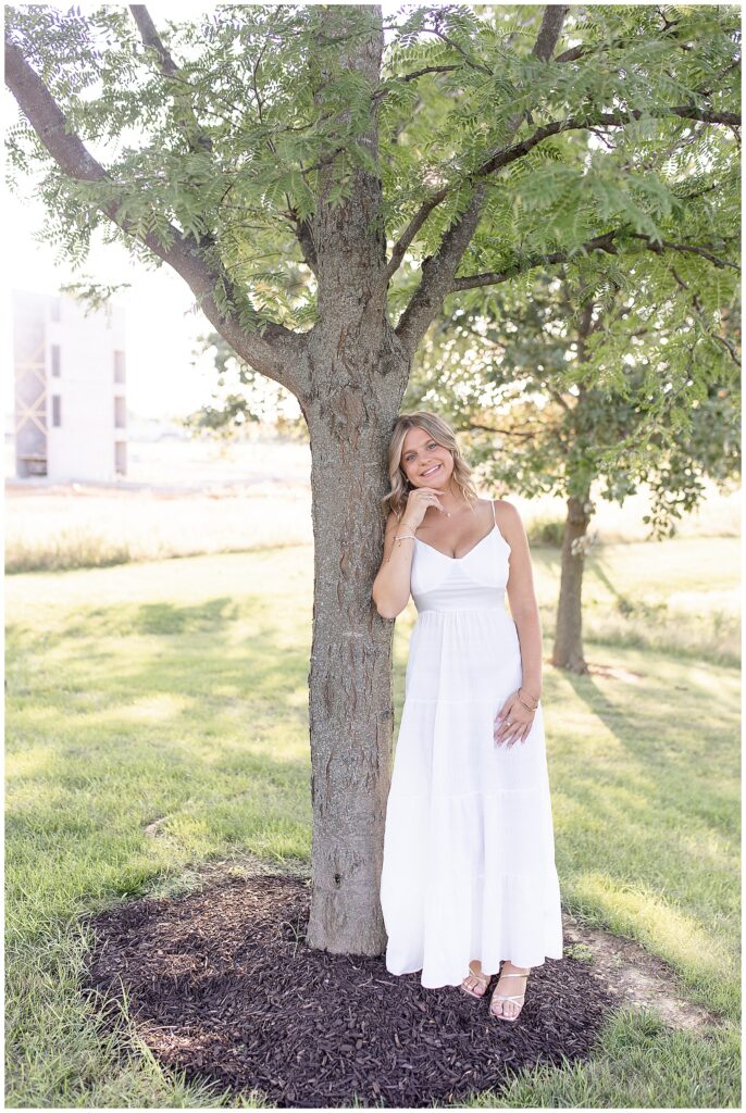 senior girl in long white dress leaning against tree at sunset at overlook park