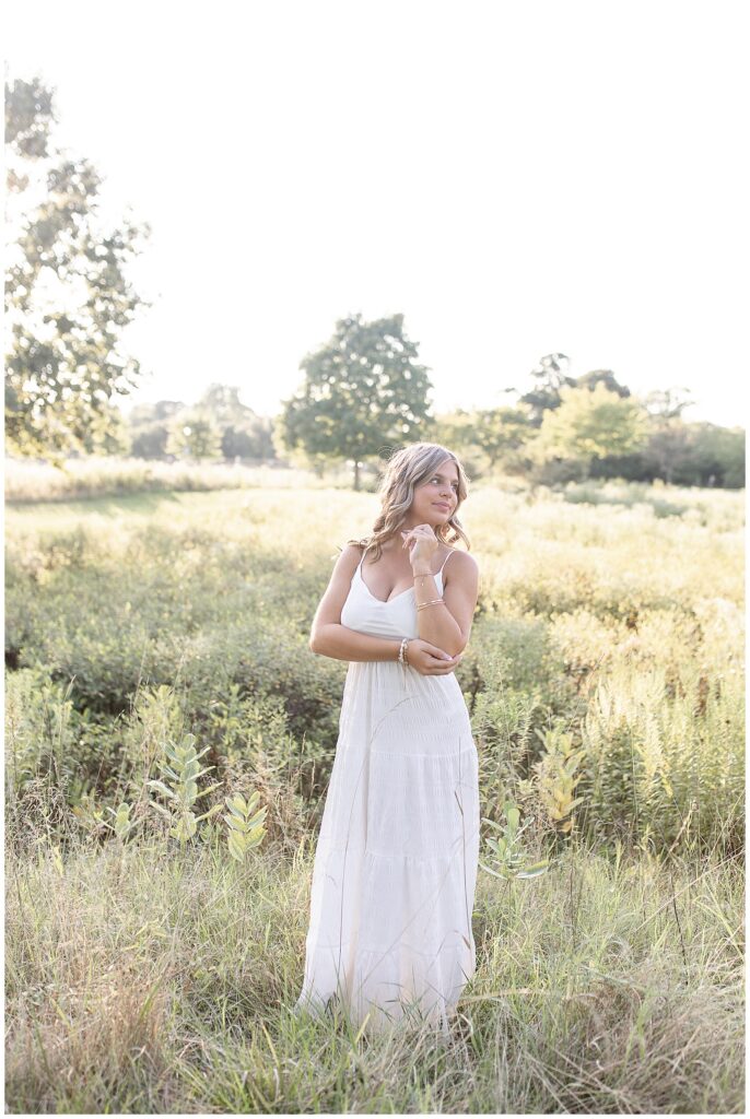 senior girl in long white dress looking over left shoulder by beautiful wildflower field at sunset in lancaster pennsylvania