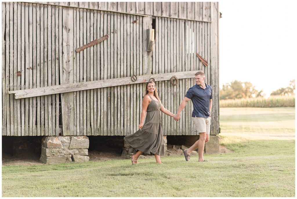 couple holding hands as guy leads girl and she looks back holding edge of her dress by barn in maryland